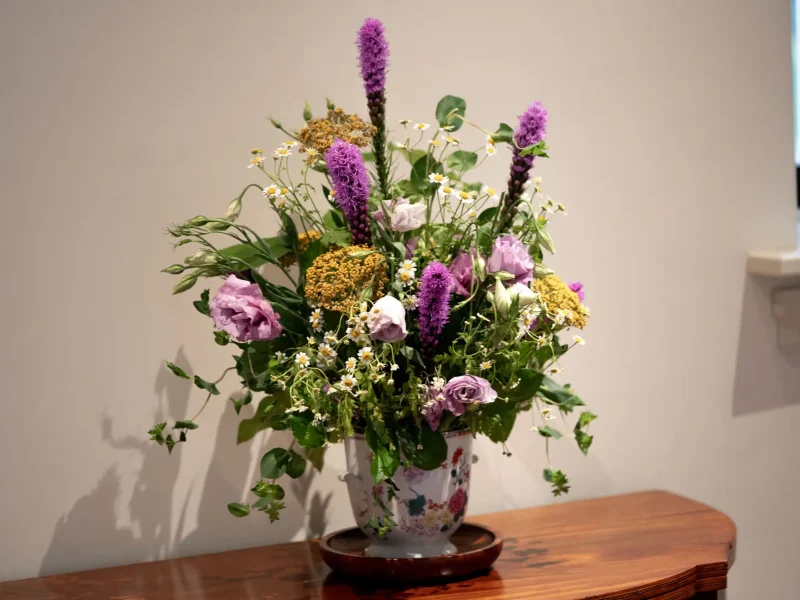 Colorful bouquet of pink, purple, and white flowers in a floral vase on a wooden table against a beige wall.